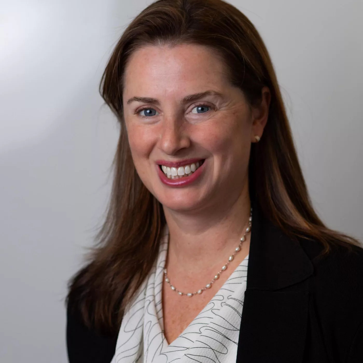 A woman wearing a black blazer and a patterned blouse is accessorized with a pearl necklace. The setting appears to be indoors with a neutral background. Her hair is styled straight, complementing the professional look.